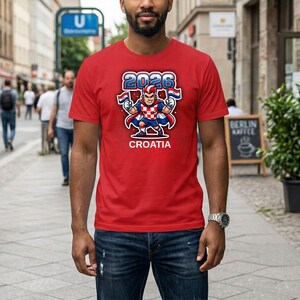Puede incluir: Una camiseta roja con una ilustraci&oacute;n de dibujos animados de un personaje sosteniendo banderas croatas, con el texto "2026" y "CROATIA". El personaje viste un atuendo con el tema de la bandera croata. La camiseta la lleva una persona.