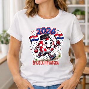 Pode incluir: Camiseta branca com um personagem de bola de futebol de desenho animado nas cores croatas, correndo em um caminho. O design inclui o ano "2026" e o texto "ŽIVJELA HRVATSKA". O personagem est&aacute; segurando duas bandeiras croatas.