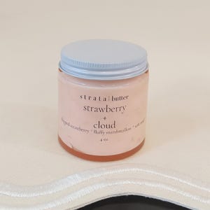Puede incluir: Un tarro de manteca corporal con aroma a fresa y nube. El tarro tiene una tapa azul claro y una etiqueta con las palabras "strata butter strawberry + cloud". El tarro contiene 118 ml de producto.