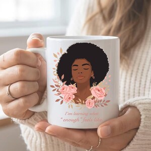 Peut inclure: Mug en céramique blanche avec une illustration d'une femme afro, des roses roses et du feuillage. Le mug porte l'inscription "I'm learning what 'enough' feels like" en rose. Le mug est tenu par une personne.