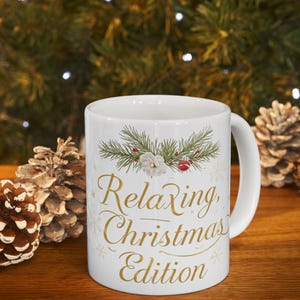 Peut inclure: Mug en céramique blanche avec l'inscription "Relaxing Christmas Edition" en lettres dorées. Le mug présente un motif festif d'aiguilles de pin, de fleurs blanches et de baies rouges. Le mug est posé sur une surface en bois avec des pommes de pin en arrière-plan.