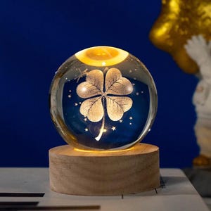 Può includere: Una sfera di vetro trasparente con un design a quadrifoglio, illuminata dall'interno, posta su una base di legno. La sfera ha uno sfondo blu con dettagli di stelle. La parte superiore è illuminata da una calda luce gialla.
