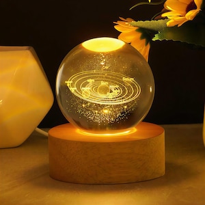 Op de afbeelding: Een gloeiende glazen bol met een verlicht zonnestelselontwerp, rustend op een houten voet. De bol toont de zon en planeten in een gedetailleerd, geëtst patroon. Het warme licht creëert een gezellige sfeer.