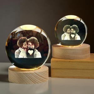 Può includere: Due sfere di cristallo illuminate con la foto di una coppia che si bacia, poste su basi di legno. Le sfere brillano di una luce calda, mettendo in risalto l'immagine incisa. Le basi in legno aggiungono un tocco naturale all'arredamento personalizzato.