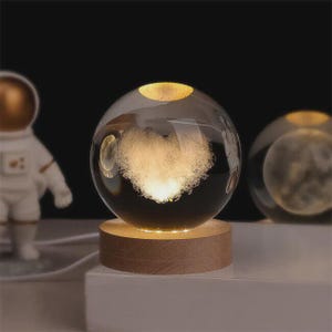 May include: A clear glass sphere with a heart-shaped design illuminated from within, resting on a wooden base. The sphere is lit with a warm glow, and a small astronaut figurine is visible in the background. The base is made of light brown wood.