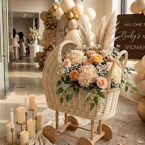 May include: A wicker baby carriage filled with a floral arrangement of roses, hydrangeas, and greenery. The carriage is on wooden wheels and stands on a beige rug. The scene is decorated with balloons and candles, suggesting a baby shower.
