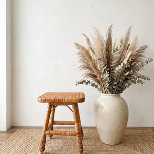 Könnte beinhalten: Ein quadratischer Hocker aus geflochtenem Rattan mit vier Beinen und einer kreuzförmigen Stützstruktur, neben einer großen, cremeweißen Keramikvase mit einem Arrangement aus getrocknetem Pampasgras und grünen Eukalyptuszweigen.