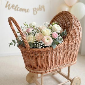 May include: A wicker baby carriage filled with a bouquet of white, pink, and peach roses, baby's breath, and eucalyptus. The carriage has a handle and is set on a wooden frame with wheels. A banner that says "Welcome Baby" is in the background.
