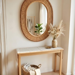 May include: An oval wall mirror with a woven rattan frame hangs above a light wood console table. On the table sits a ceramic vase with dried flowers and a small dish. A woven basket containing books and a folded textile rests on the floor below.