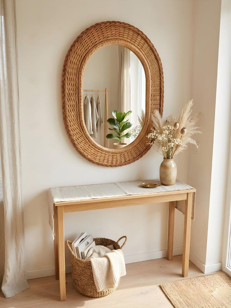 May include: An oval wall mirror with a woven rattan frame hangs above a light wood console table. On the table sits a ceramic vase with dried flowers and a small dish. A woven basket containing books and a folded textile rests on the floor below.