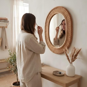 May include: An oval wall mirror with a woven light brown rattan frame. The mirror is mounted on a plain white wall above a light wood console table. The frame features a detailed braided texture around the circular glass surface.