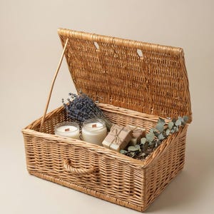 Puede incluir: Una cesta rectangular de mimbre trenzado con tapa abatible abierta y asa frontal. Dentro de la cesta hay dos velas de cristal con mechas de madera, dos pastillas de jabón rectangulares envueltas en papel marrón con cordel, lavanda y eucalipto.