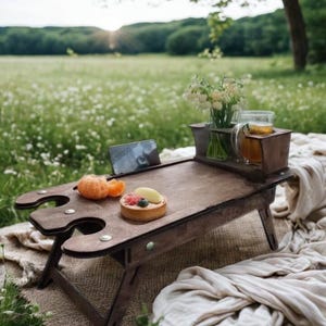 May include: A dark brown wooden picnic table with a built-in wine glass holder and a tablet stand. The table holds a tart, orange slices, and a glass of tea. The table is set on a blanket in a field of wildflowers.