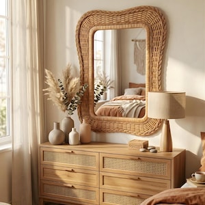 May include: A rectangular wall mirror with a thick, wavy woven rattan frame. Below the mirror sits a light wood dresser featuring six drawers with cane webbing panels, topped with four ceramic vases, dried grasses, and a table lamp.