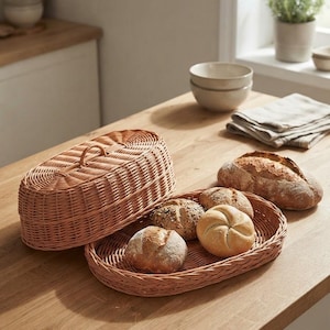 May include: An oval-shaped light brown woven wicker bread basket with a matching removable lid. The basket contains three small round bread rolls, while a larger loaf of crusty artisan bread sits separately on the surface next to the basket.