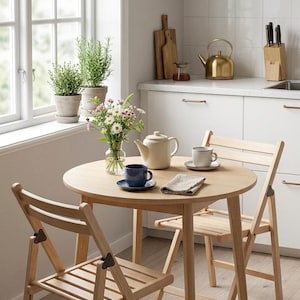 Puede incluir: Una mesa de comedor redonda de madera clara con dos sillas plegables a juego. Sobre la mesa hay una tetera de color crema, una taza azul sobre un platillo, una taza blanca sobre un platillo, una servilleta doblada y un florero de cristal.