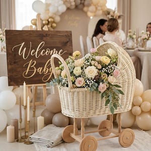May include: A wicker baby carriage filled with flowers, set against a backdrop of balloons and a wooden sign that reads "Welcome Baby." The carriage has wooden wheels and is filled with roses and other flowers.