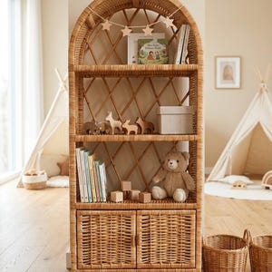 Puede incluir: Una estantería alta de ratán con parte superior arqueada, diseño de celosía abierta, tres estantes abiertos y un armario inferior con dos puertas tejidas. Los estantes contienen libros, figuras de madera, una caja y un oso de peluche.