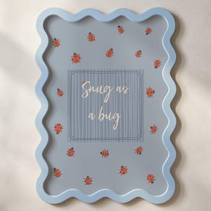 May include: A light blue, wavy-edged picture frame with a light blue background. The frame features scattered red ladybug illustrations and the text "Snug as a bug" in the center.