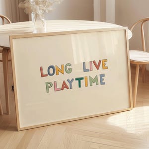 Può includere: Una stampa artistica incorniciata con le parole "LONG LIVE PLAYTIME" in lettere a blocchi colorate. L'opera d'arte è in una cornice di legno chiaro ed è esposta su una superficie di colore chiaro. La stampa è circondata da un tavolo e sedie in legno chiaro.