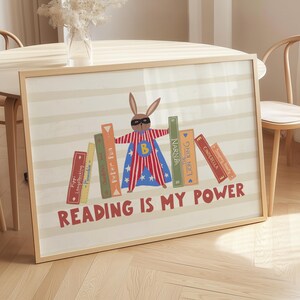 Op de afbeelding: Een ingelijste print met een superheld konijn met masker en cape, voor kleurrijke boeken. De tekst "READING IS MY POWER" staat in het rood. De achtergrond heeft beige en witte strepen. Kinderkamer decoratie.