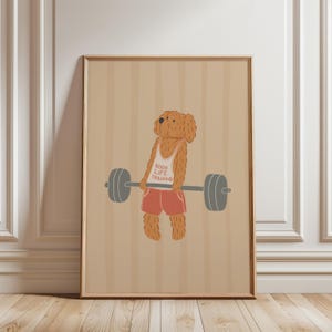 Op de afbeelding: Een ingelijste prent met een cartoonhond die gewichten heft. De bruine hond draagt een witte tanktop met "GOOD LIFE TRAINING" en een rode korte broek. De achtergrond is beige met verticale strepen.