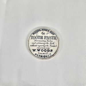Puede incluir: Una lata circular vintage de "WOODS ARECA NUT TOOTH PASTE". La lata blanca tiene texto negro que dice "Para eliminar el sarro y blanquear los dientes sin dañar el esmalte". El propietario es W. WOODS, Chemist, Plymouth.