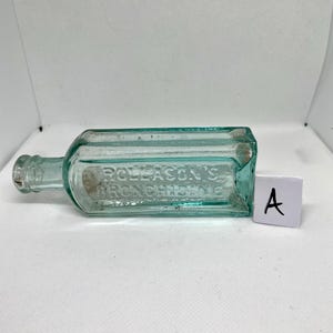 K&ouml;nnte beinhalten: Eine rechteckige, hellgr&uuml;ne Glasflasche im Vintage-Stil mit dem gepr&auml;gten Text "ROLLASON'S BRONCHITIS". Die Flasche hat einen schmalen Hals und ein kleines Etikett. Ein wei&szlig;es Etikett mit dem Buchstaben "A" befindet sich neben der Flasche.