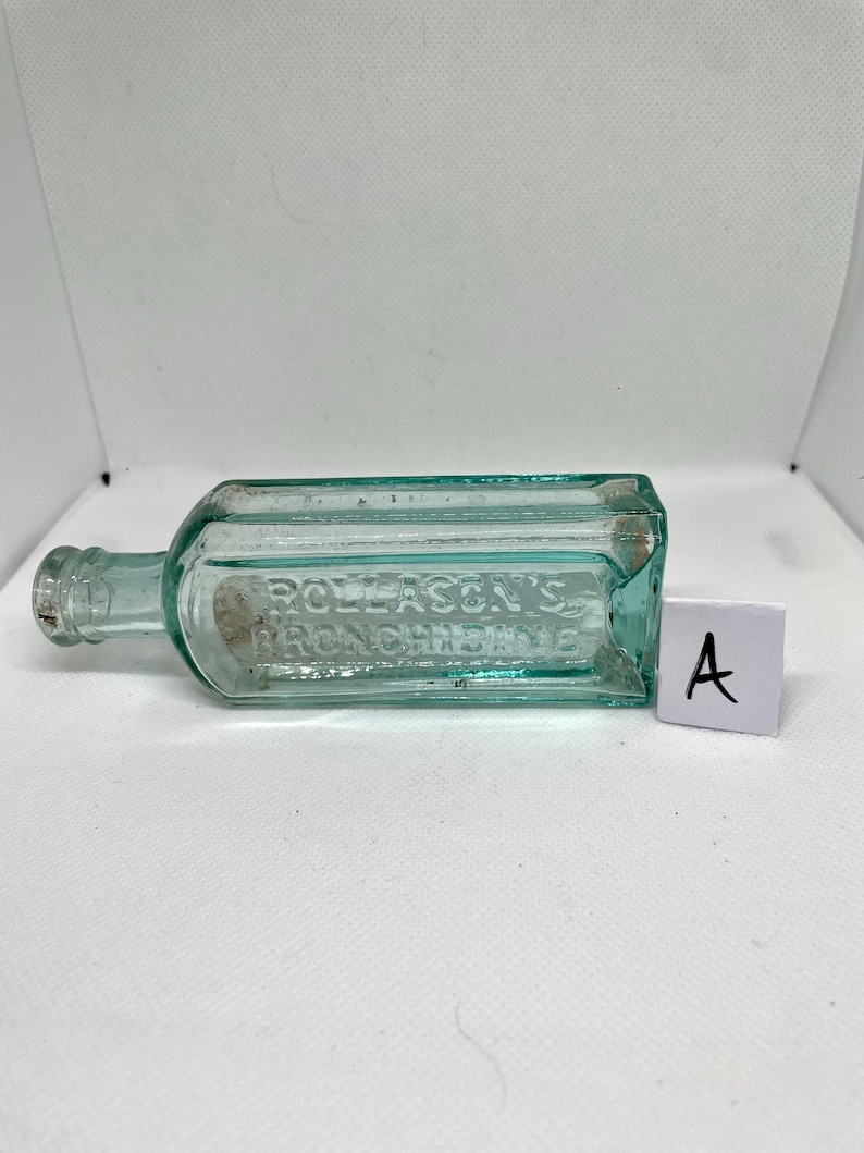 K&ouml;nnte beinhalten: Eine rechteckige, hellgr&uuml;ne Glasflasche im Vintage-Stil mit dem gepr&auml;gten Text "ROLLASON'S BRONCHITIS". Die Flasche hat einen schmalen Hals und ein kleines Etikett. Ein wei&szlig;es Etikett mit dem Buchstaben "A" befindet sich neben der Flasche.