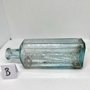 K&ouml;nnte beinhalten: Eine rechteckige, hellblaue antike Glasflasche mit rundem Hals und kleiner Lippe. Die Flasche hat einen gepr&auml;gten Text und liegt auf einer wei&szlig;en Oberfl&auml;che. Eine kleine wei&szlig;e Karte mit dem Buchstaben "B" befindet sich im Vordergrund.