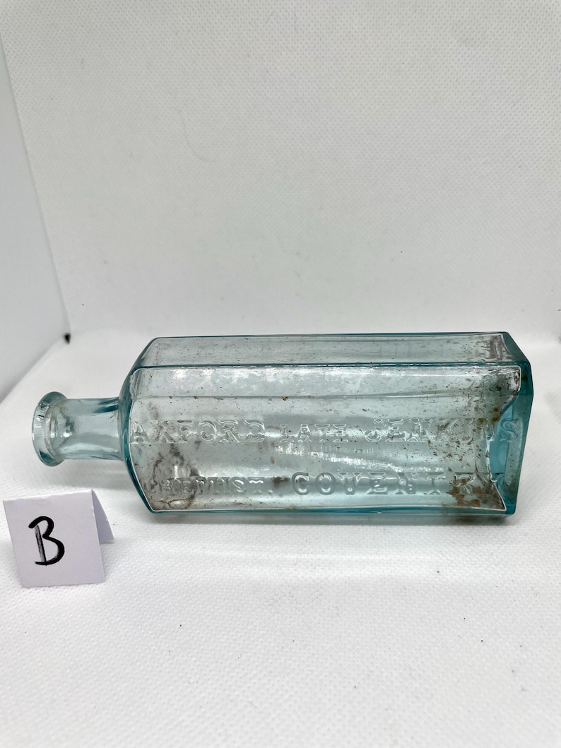 K&ouml;nnte beinhalten: Eine rechteckige, hellblaue antike Glasflasche mit rundem Hals und kleiner Lippe. Die Flasche hat einen gepr&auml;gten Text und liegt auf einer wei&szlig;en Oberfl&auml;che. Eine kleine wei&szlig;e Karte mit dem Buchstaben "B" befindet sich im Vordergrund.