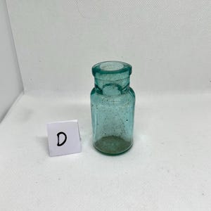 K&ouml;nnte beinhalten: Kleine, antike, teal-farbene Glasflasche mit zylindrischem K&ouml;rper und rundem Boden. Die Flasche hat einen kurzen Hals und eine weite &Ouml;ffnung. Ein wei&szlig;es Quadrat mit dem Buchstaben "D" befindet sich daneben.
