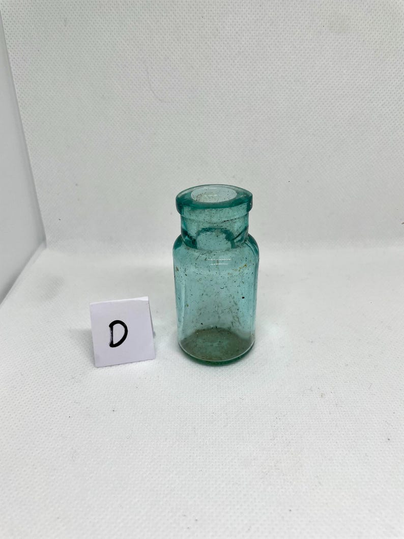 K&ouml;nnte beinhalten: Kleine, antike, teal-farbene Glasflasche mit zylindrischem K&ouml;rper und rundem Boden. Die Flasche hat einen kurzen Hals und eine weite &Ouml;ffnung. Ein wei&szlig;es Quadrat mit dem Buchstaben "D" befindet sich daneben.
