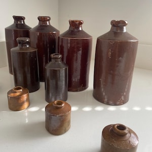 May include: A selection of antique brown stoneware bottles and jars. The bottles have a glossy finish and rounded shoulders, with a small opening at the top. The jars are cylindrical with a similar brown glaze. The collection includes both tall and short vessels.