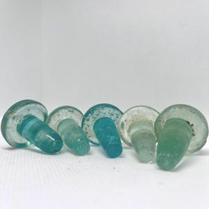Peut inclure: Ensemble de cinq bouchons de bouteille en verre, dans des tons de bleu et de vert. Chaque bouchon a un dessus arrondi et une base effilée. Le verre est texturé et translucide, avec des variations de couleur.
