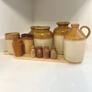 Puede incluir: Colección de tarros y botellas antiguos de gres en varios tamaños y formas. Las piezas son principalmente de color marrón y beige, con un diseño de dos tonos. Los artículos están dispuestos sobre una bandeja de madera, mostrando su estética vintage.