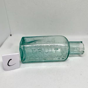 K&ouml;nnte beinhalten: Eine rechteckige, teal-farbene Glasflasche mit dem gepr&auml;gten Text "FRUIT CRYSTALS". Die Flasche hat einen kurzen Hals und eine leicht besch&auml;digte &Ouml;ffnung. Neben der Flasche befindet sich ein kleines wei&szlig;es Quadrat mit dem Buchstaben "C".