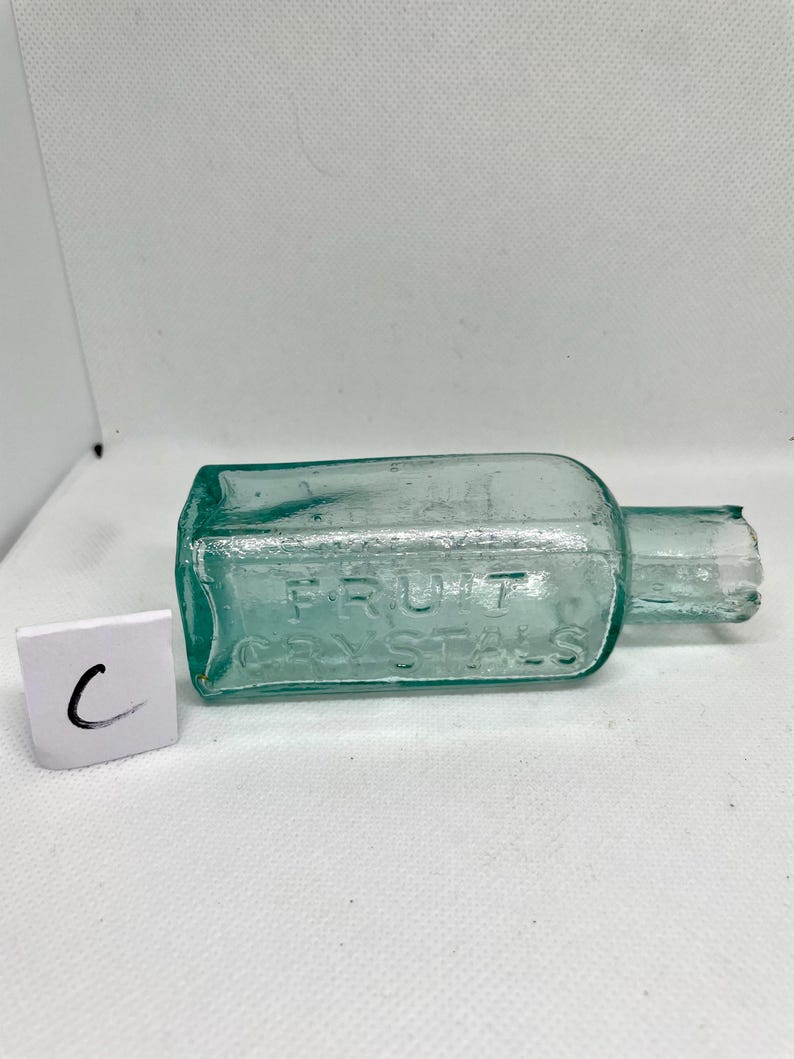 K&ouml;nnte beinhalten: Eine rechteckige, teal-farbene Glasflasche mit dem gepr&auml;gten Text "FRUIT CRYSTALS". Die Flasche hat einen kurzen Hals und eine leicht besch&auml;digte &Ouml;ffnung. Neben der Flasche befindet sich ein kleines wei&szlig;es Quadrat mit dem Buchstaben "C".