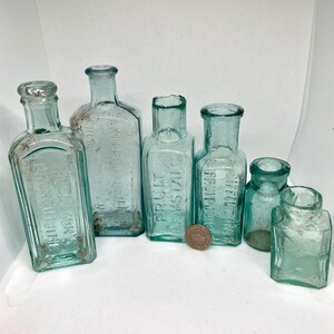 K&ouml;nnte beinhalten: Eine Sammlung antiker, hellblauer Glasflaschen in verschiedenen Formen und Gr&ouml;&szlig;en. Einige Flaschen haben gepr&auml;gten Text, darunter "FRUIT CRYSTAL" und "RULLASON'S RHEUMATIDINE". Eine M&uuml;nze dient als Ma&szlig;stab.