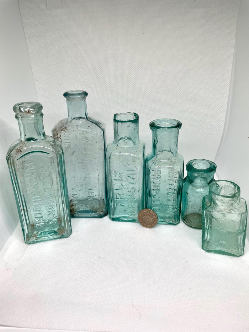 K&ouml;nnte beinhalten: Eine Sammlung antiker, hellblauer Glasflaschen in verschiedenen Formen und Gr&ouml;&szlig;en. Einige Flaschen haben gepr&auml;gten Text, darunter "FRUIT CRYSTAL" und "RULLASON'S RHEUMATIDINE". Eine M&uuml;nze dient als Ma&szlig;stab.
