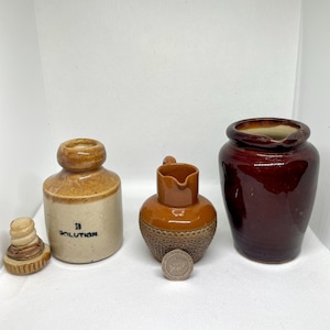 May include: A collection of antique ceramic vessels and a cork stopper. The items include a small brown and tan bottle with the word "SOLUTION", a small brown pitcher, and a larger maroon jar.