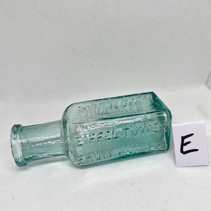 K&ouml;nnte beinhalten: Eine antike, rechteckige, hellgr&uuml;ne Glasflasche mit dem gepr&auml;gten Text "EIFFEL TOWER FRUIT JUICES". Die Flasche hat einen kurzen Hals und eine kleine &Ouml;ffnung. Ein wei&szlig;es Quadrat mit dem Buchstaben "E" befindet sich neben der Flasche.