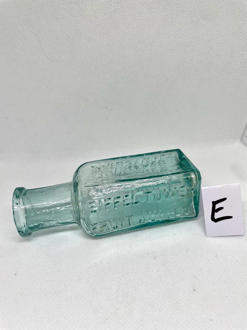 K&ouml;nnte beinhalten: Eine antike, rechteckige, hellgr&uuml;ne Glasflasche mit dem gepr&auml;gten Text "EIFFEL TOWER FRUIT JUICES". Die Flasche hat einen kurzen Hals und eine kleine &Ouml;ffnung. Ein wei&szlig;es Quadrat mit dem Buchstaben "E" befindet sich neben der Flasche.