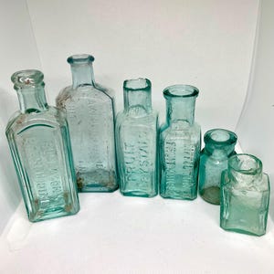 K&ouml;nnte beinhalten: Eine Sammlung antiker Glasflaschen in verschiedenen Formen und Gr&ouml;&szlig;en. Die Flaschen sind hellblaugr&uuml;n und scheinen mit Text gepr&auml;gt zu sein. Einige Flaschen haben sichtbare Etiketten, darunter "Fruit Crystals" und "Rollason's Perchloridine".