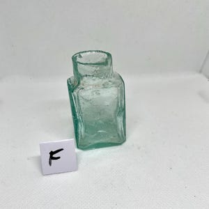 K&ouml;nnte beinhalten: Kleine, antike, quadratische Glasflasche mit leicht gr&uuml;nlicher T&ouml;nung. Die Flasche hat einen kurzen Hals und eine leicht unebene Oberfl&auml;che. Ein kleines wei&szlig;es Quadrat mit dem Buchstaben "F" befindet sich vor der Flasche.