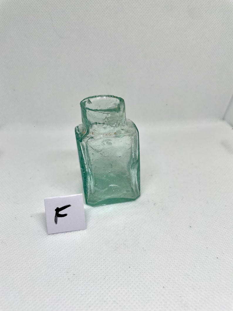 K&ouml;nnte beinhalten: Kleine, antike, quadratische Glasflasche mit leicht gr&uuml;nlicher T&ouml;nung. Die Flasche hat einen kurzen Hals und eine leicht unebene Oberfl&auml;che. Ein kleines wei&szlig;es Quadrat mit dem Buchstaben "F" befindet sich vor der Flasche.