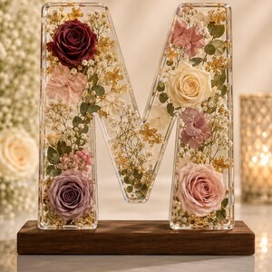 May include: A clear resin letter M containing preserved flowers including a dark red rose, a light pink rose, a cream rose, and a lavender rose, accented with white baby's breath and gold leaf flakes, mounted on a rectangular dark wood base.