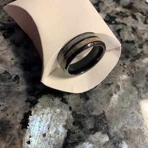 May include: A black ring with a silver and tan band, presented in a white paper holder. The ring has a polished finish and a modern design. The ring is likely made of metal and wood.