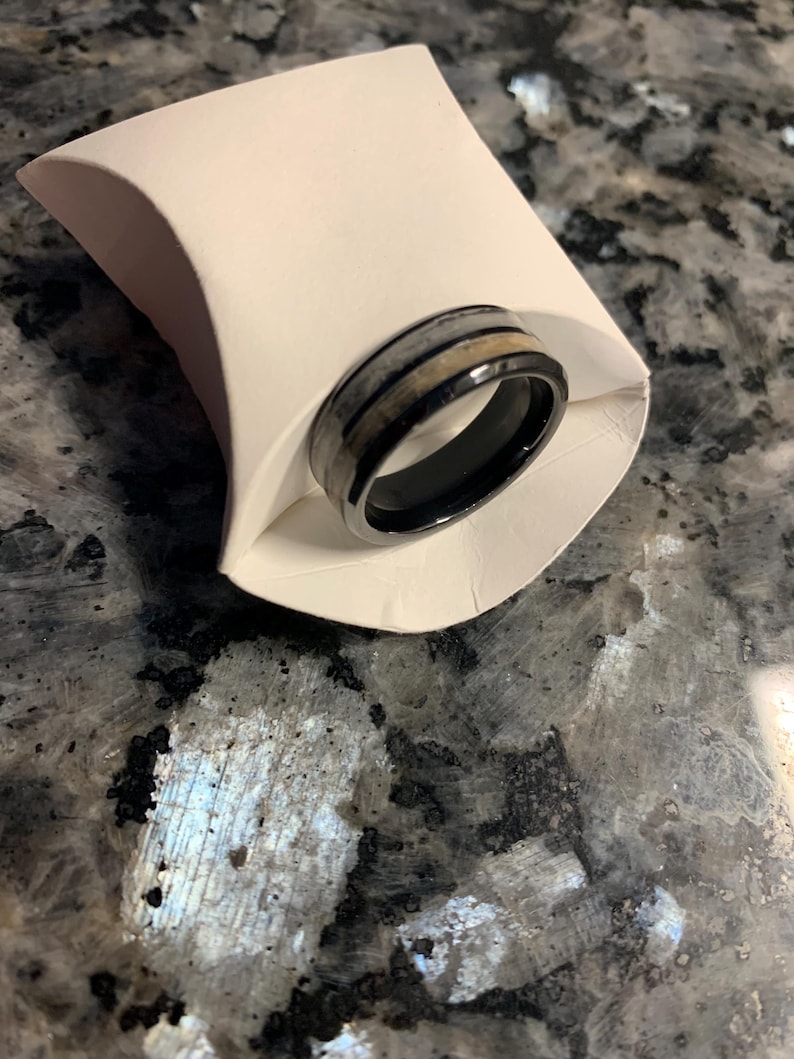May include: A black ring with a silver and tan band, presented in a white paper holder. The ring has a polished finish and a modern design. The ring is likely made of metal and wood.