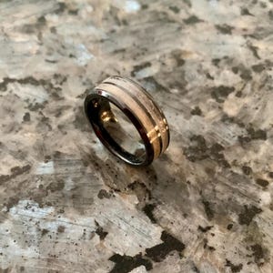 May include: A black ring with a wooden inlay and a silver band. The ring has a unique design with alternating light and dark wood grain patterns. The ring is likely made of tungsten carbide and wood.